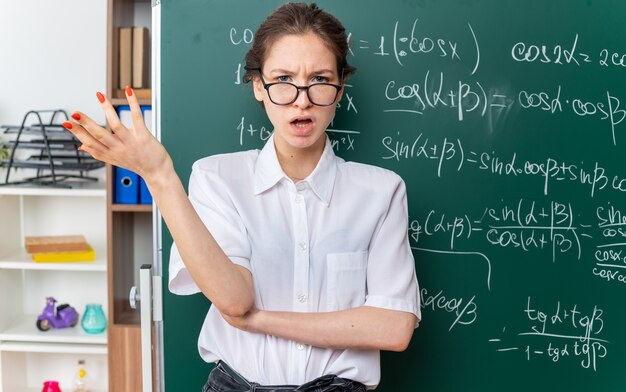 Verärgerte junge Mathematiklehrerin mit Brille, die vor der Tafel steht und die leere Hand zeigt, die im Klassenzimmer nach vorne schaut