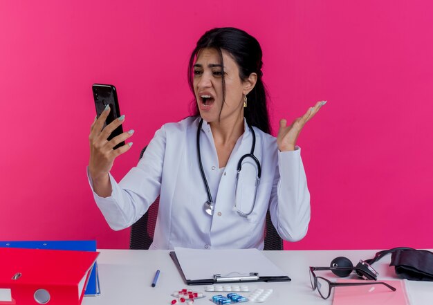 Verärgerte junge Ärztin, die medizinische Robe und Stethoskop trägt, sitzt am Schreibtisch mit medizinischen Werkzeugen, die Handy halten und betrachten und leere Hand lokalisiert auf rosa Wand zeigen