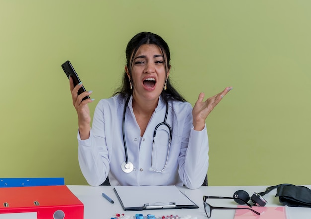 Verärgerte junge Ärztin, die medizinische Robe und Stethoskop trägt, sitzt am Schreibtisch mit medizinischen Werkzeugen, die Handy suchen, das leere Hand lokalisiert zeigt