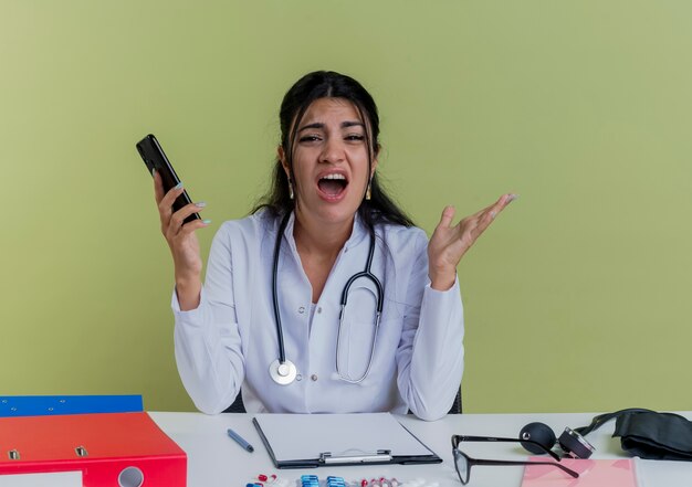 Verärgerte junge Ärztin, die medizinische Robe und Stethoskop trägt, sitzt am Schreibtisch mit medizinischen Werkzeugen, die Handy suchen, das leere Hand lokalisiert zeigt