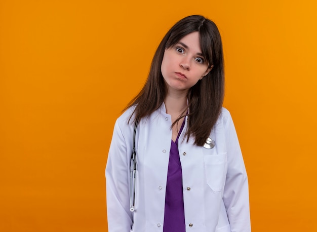 Verärgerte junge Ärztin im medizinischen Gewand mit Stethoskop betrachtet Kamera auf lokalisiertem orangefarbenem Hintergrund mit Kopienraum