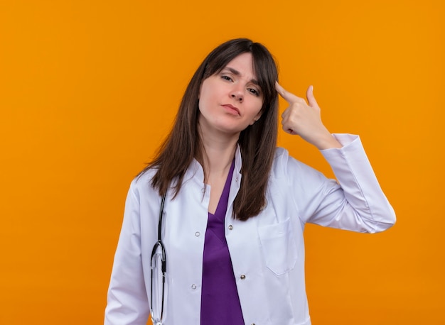Verärgerte junge Ärztin im medizinischen Gewand mit Stethoskop gestikuliert Kopfschuss mit Hand auf lokalisiertem orangefarbenem Hintergrund mit Kopienraum