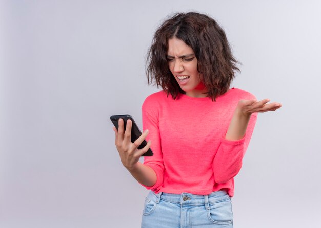 Verärgerte junge schöne Frau, die Handy hält und leere Hand auf isolierter weißer Wand mit Kopienraum zeigt