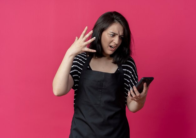Verärgerte junge schöne weibliche Friseurin in der Schürze, die Bildschirm ihres Handys mit genervtem Ausdruck über Rosa betrachtet