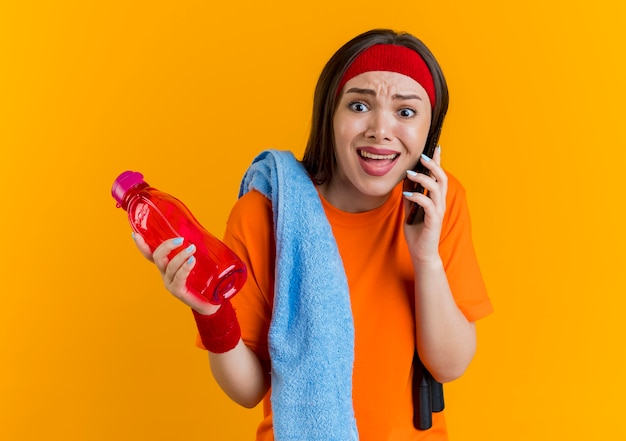 Verärgerte junge sportliche Frau, die Stirnband und Armbänder mit Springseil und Handtuch auf Schultern trägt Wasserflasche hält und am Telefon gerade schaut