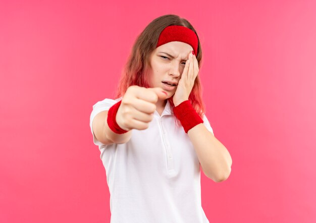 Verärgerte junge sportliche Frau im Stirnband, die ihr Auge berührt, zeigt Faust zur Kamera mit unglücklichem Gesicht, das über rosa Wand steht