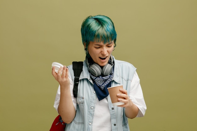 Verärgerte junge Studentin mit Kopfhörern und Kopftuch am Hals und Rucksack mit Kaffeetasse aus Papier und Kappe, die isoliert auf olivgrünem Hintergrund in die Tasse schaut