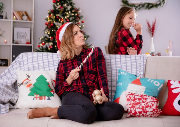 Verärgerte Mutter mit Weihnachtsmütze hält einen Teil der zerbrochenen Zuckerstange, die auf der Couch sitzt, und schaut auf erfreute Tochter, die Zuckerstange isst, die Weihnachtszeit zu Hause genießt