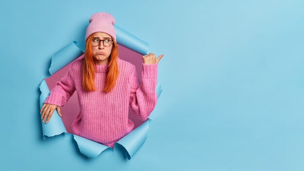 Kostenloses Foto verärgerte niedergeschlagene frau mit roten haaren, die in niederem geist sind und mit unzufriedenem ausdruck auf kopierraum zeigen