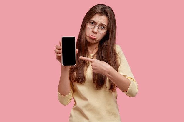 Verärgerte niedergeschlagene Frau spitzt die Unterlippe, zeigt auf ein modernes Gerät, zeigt einen leeren Bildschirm für Ihren Text, mag nicht, wie es funktioniert, trägt eine runde Brille