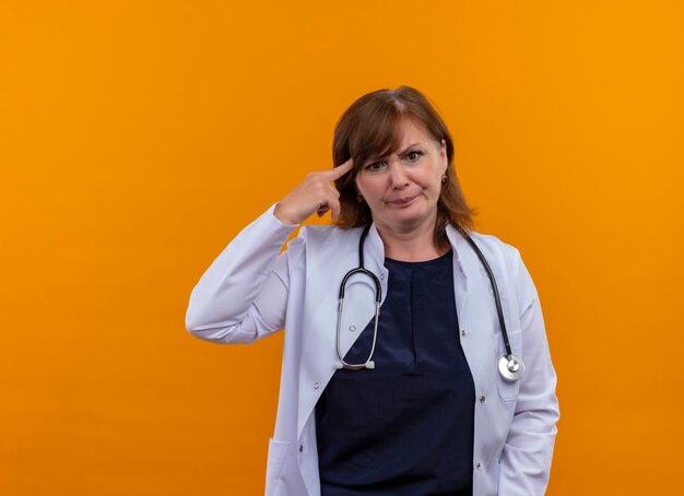 Verärgerte Ärztin mittleren Alters, die medizinische Robe und Stethoskop trägt, die Finger auf Schläfe auf isolierte orange Wand mit Kopienraum setzen