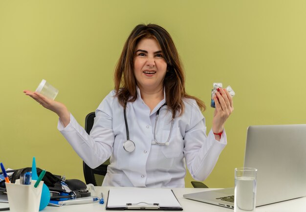 Verärgerte Ärztin mittleren Alters, die medizinische Robe und Stethoskop trägt, sitzt am Schreibtisch mit medizinischen Werkzeugen und Laptop, die medizinische Drogen und Becher isoliert halten