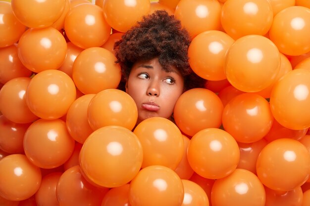 Verärgerte traurige Frau mit Afro-Haaren ragt Kopf durch aufgeblasene Luftballons heraus sieht traurig weg, will nicht alt werden, umgeben von orangefarbenen Heliumballons, die auf Geburtstagsfeier einsam sind