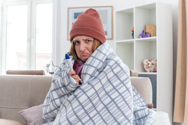 Kostenloses Foto verärgerte ungesunde junge frau in warmem hut, eingewickelt in decke, die unwohl und krank aussieht und an erkältung und grippe leidet, mit thermometer mit fieber, das besorgt auf der couch im hellen wohnzimmer sitzt