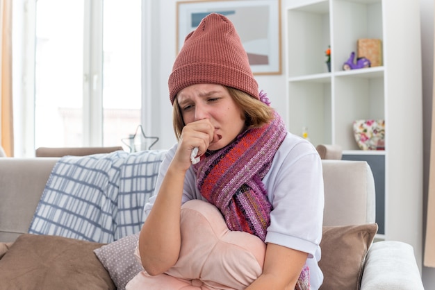 Verärgerte ungesunde junge Frau in warmem Hut mit Schal, die unwohl aussieht und krankes Gewebe hält, das an Erkältung und Grippe leidet und Nase bläst, die auf der Couch im hellen Wohnzimmer sitzt