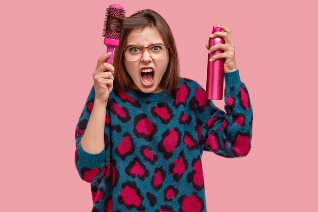 Verärgerte unzufriedene weibliche Kämme Haare, empört, macht Frisur mit Haarbürste und Spray