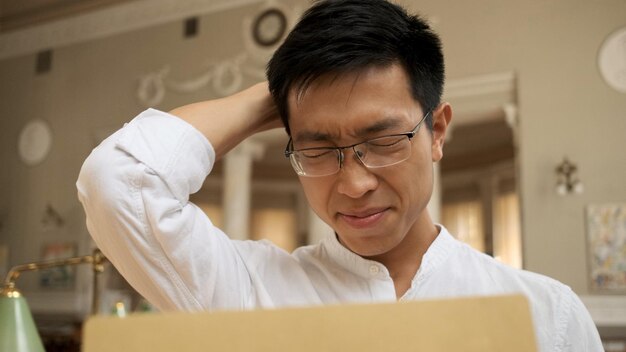 Verärgerter asiatischer männlicher Student, der Umschlag mit Prüfungsergebnissen in der Bibliothek öffnet und traurig den Kopf hält