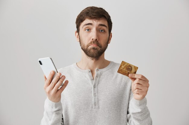 Verärgerter bärtiger Kerl mit Kreditkarte und Smartphone