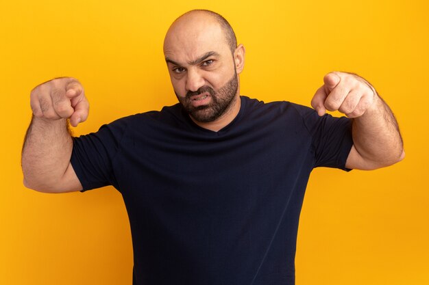 Verärgerter bärtiger Mann im Marine-T-Shirt, das mit Zeigefingern zeigt, die über orange Wand stehen