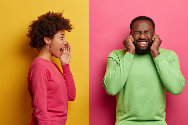 Verärgerter bärtiger Mann steckt die Ohren zu, beißt die Zähne zusammen, will die Frau nicht schreien hören, trägt einen grünen Pullover. Afroamerikanerin hält Handfläche in der Nähe des Mundes, schreit und sieht Ehemann an, steht im Profil