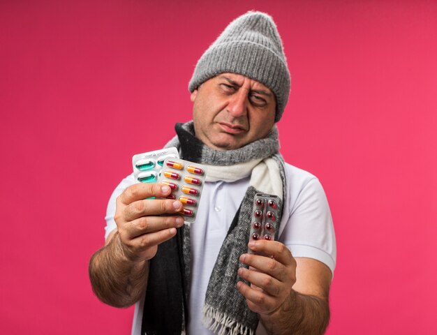 verärgerter erwachsener kranker kaukasischer Mann mit Schal um den Hals, der eine Wintermütze trägt, die verschiedene Medizinpackungen isoliert auf rosa Wand mit Kopienraum hält