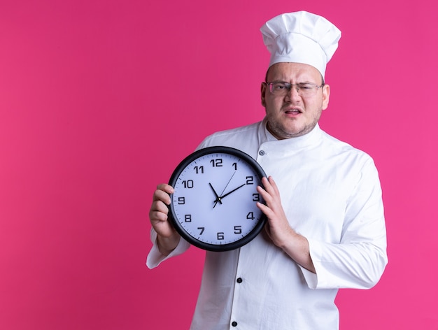 Verärgerter erwachsener männlicher Koch, der Kochuniform und Brille trägt, die die Uhr mit Blick auf die Vorderseite isoliert auf rosa Wand mit Kopienraum hält