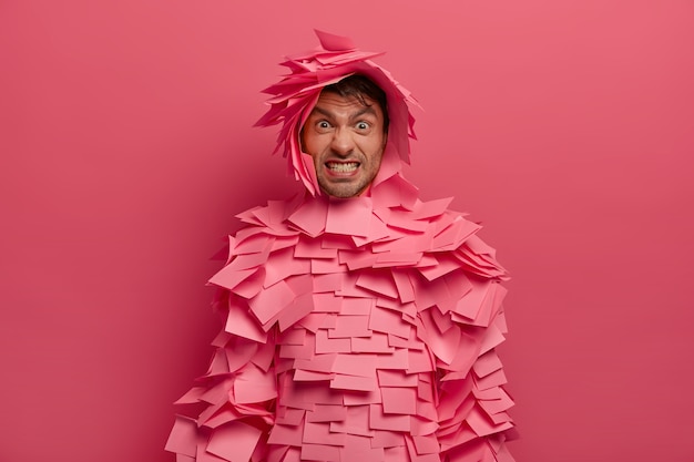 Verärgerter erwachsener Mann beißt die Zähne zusammen, macht eine wütende Grimasse, bedeckt mit Haftnotizen, hat ein kreatives Kostüm, biss die Zähne zusammen, isoliert über der rosa Wand, runzelt die Stirn. Konzept des menschlichen Gesichtsausdrucks