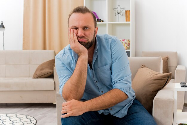 Verärgerter erwachsener slawischer Mann sitzt auf Sessel, der Hand auf Gesicht setzt und Kamera im Wohnzimmer betrachtet