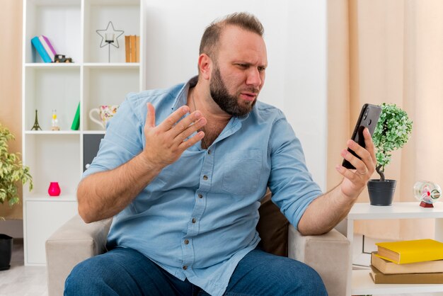 Verärgerter erwachsener slawischer Mann sitzt auf Sessel mit erhobener Hand und schaut auf Telefon im Wohnzimmer