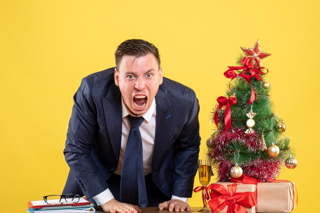 Verärgerter Geschäftsmann der Vorderansicht, der nahe Weihnachtsbaum steht und auf gelbem Hintergrund präsentiert