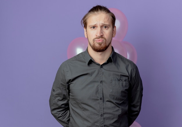 Verärgerter gutaussehender Mann hält Heliumballons hinter sich und sieht auf lila Wand isoliert aus