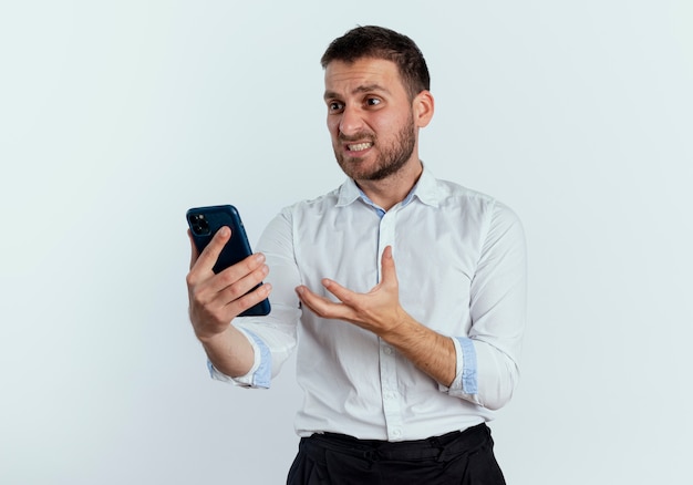 Verärgerter gutaussehender Mann hält und betrachtet Telefon, das Hand lokalisiert auf weißer Wand hebt