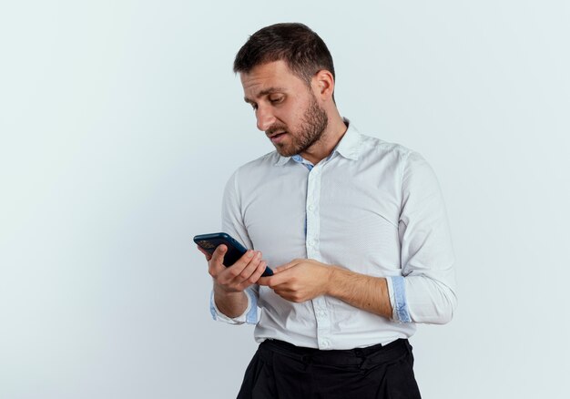 Verärgerter gutaussehender Mann hält und betrachtet Telefon lokalisiert auf weißer Wand