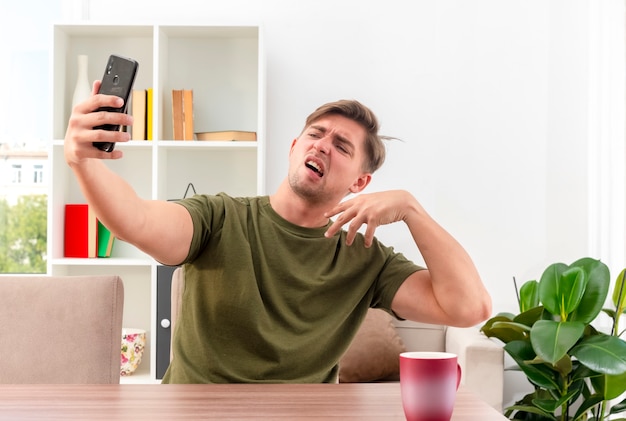 Verärgerter junger blonder gutaussehender Mann sitzt am Tisch mit der Tasse, die Hand hebt und Telefon im Wohnzimmer betrachtet