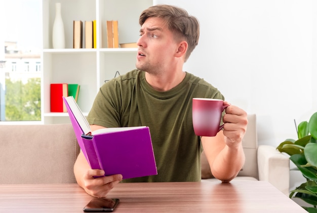 Kostenloses Foto verärgerter junger blonder gutaussehender mann sitzt am tisch mit telefon, das buch und tasse hält, die seite innerhalb des wohnzimmers betrachten