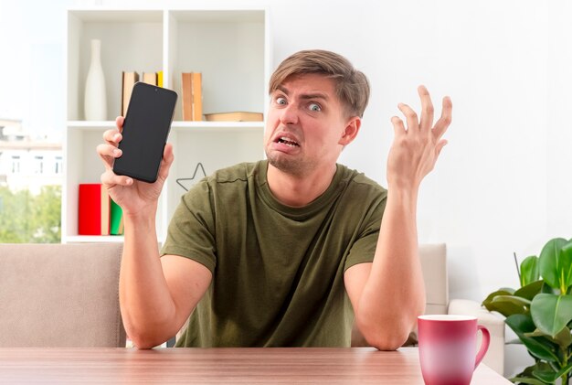 Verärgerter junger blonder gutaussehender Mann sitzt am Tisch und hebt die Hand und hält das Telefon im Wohnzimmer