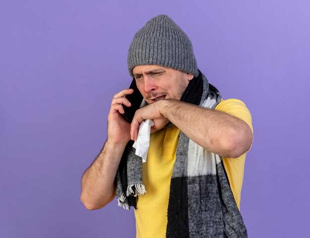 Verärgerter junger blonder kranker slawischer Mann, der Wintermütze und Schal trägt, hält Taschentuch, das am Telefon spricht