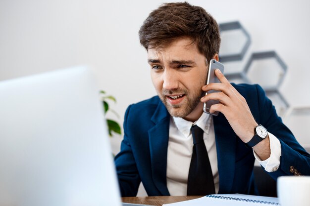 Verärgerter junger Geschäftsmann, der am Telefon, Bürohintergrund spricht.