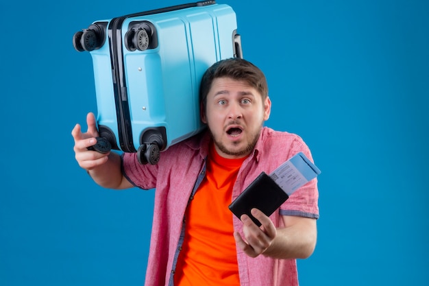 Verärgerter junger gutaussehender Reisender Mann, der Koffer und Flugtickets hält, die verwirrt sehr emotional und besorgt über der blauen Wand stehen
