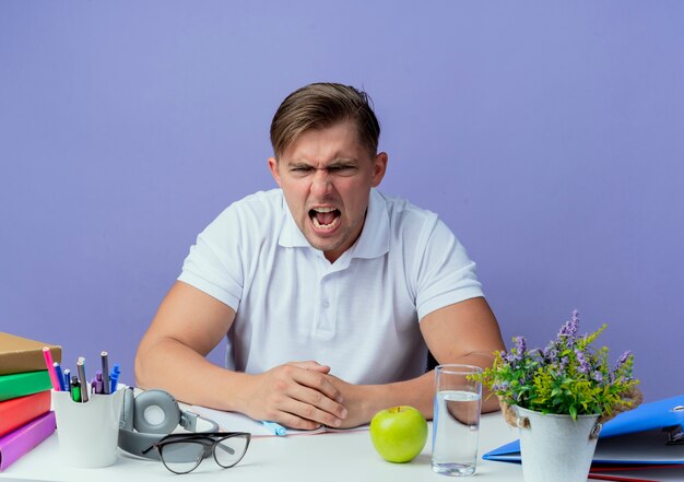 Verärgerter junger hübscher männlicher Student, der am Schreibtisch mit Schulwerkzeugen lokalisiert auf blauem Hintergrund sitzt