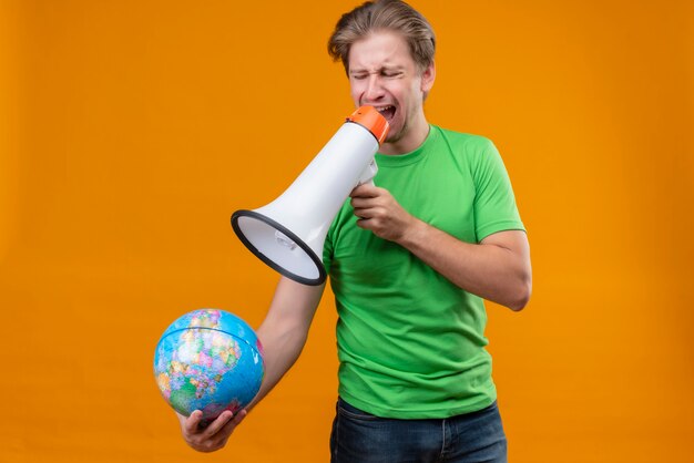 Verärgerter junger hübscher Mann, der grünes T-Shirt hält, das Globus hält, der zum Megaphon schreit, das über orange Wand steht