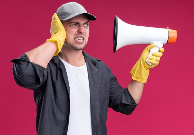 Verärgerter junger hübscher Reinigungsmann, der T-Shirt und Kappe mit Handschuhen hält, die Lautsprecher halten und betrachten, der Hand auf Kopf lokalisiert auf rosa Wand setzt