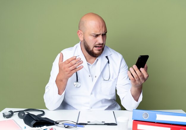 Verärgerter junger kahlköpfiger männlicher Arzt, der medizinische Robe und Stethoskop trägt, die am Schreibtisch arbeiten, mit medizinischen Werkzeugen halten und Telefon lokalisiert auf grünem Hintergrund betrachten