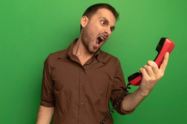 Verärgerter junger kaukasischer Mann, der altes Telefon hält und es schreiend isoliert auf grüner Wand betrachtet