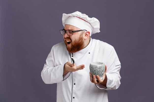 Verärgerter junger Koch mit einheitlicher Brille und Mütze mit Löffel und Schüssel, der auf die Seite zeigt, die auf die Schüssel zeigt, die auf violettem Hintergrund isoliert ist
