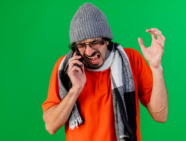 Verärgerter junger kranker Mann, der Brille Wintermütze und Schal trägt, die Hand in der Luft hält, die am Telefon mit geschlossenen Augen spricht, die auf grüner Wand lokalisiert werden