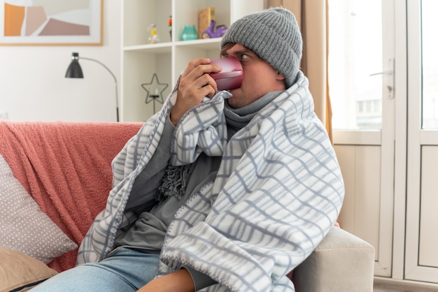 Verärgerter junger kranker Mann mit Schal um den Hals, der eine Wintermütze trägt, die in Plaid gehüllt ist und aus einer Tasse auf der Couch im Wohnzimmer trinkt?