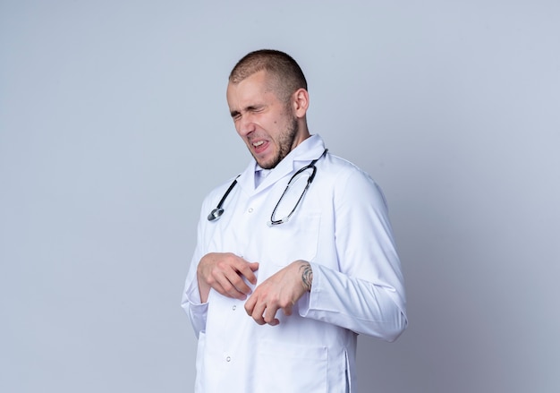Verärgerter junger männlicher Arzt, der medizinische Robe und Stethoskop um seinen Hals trägt und Hand auf Bauch mit geschlossenen Augen legt, die auf Weiß mit Kopierraum isoliert sind