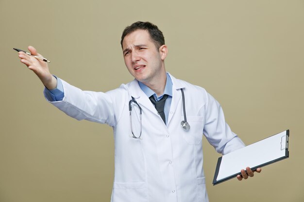 Verärgerter junger männlicher Arzt mit medizinischem Gewand und Stethoskop um den Hals, der Klemmbrett und Stift hält und auf die Seite zeigt, die mit der Hand isoliert auf olivgrünem Hintergrund zeigt