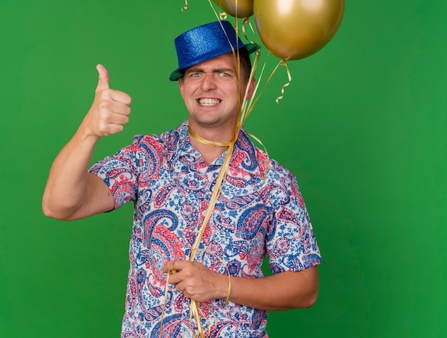 Verärgerter junger Party-Typ, der blauen Hut trägt, der Luftballons hält, die um Hals gebunden sind, zeigt Daumen oben isoliert auf Grün